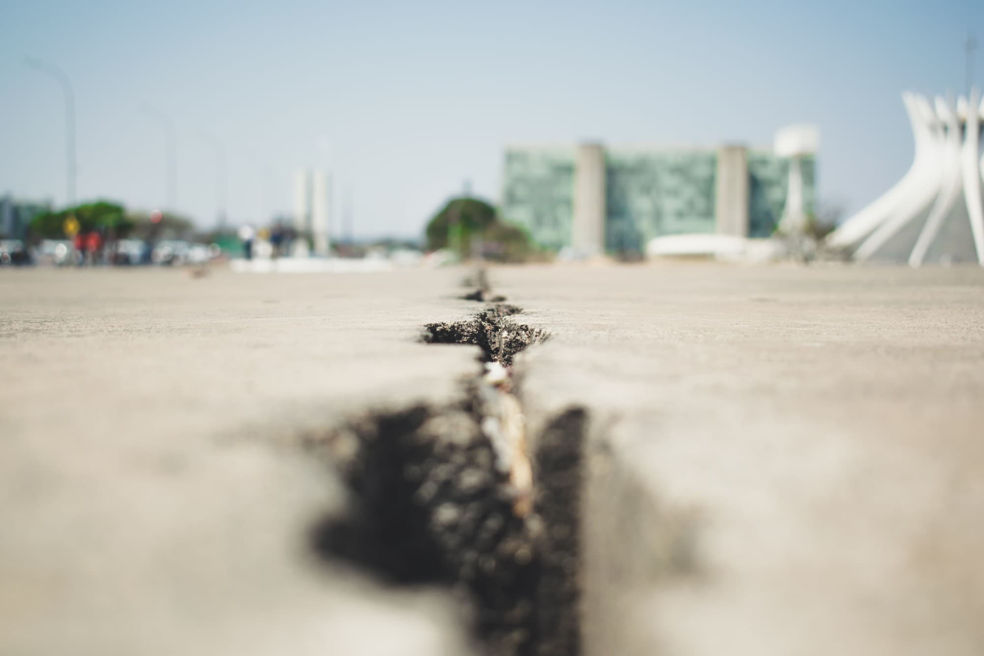 Close-up of a deep crack running through a concrete plaza, with modern buildings blurred in the background